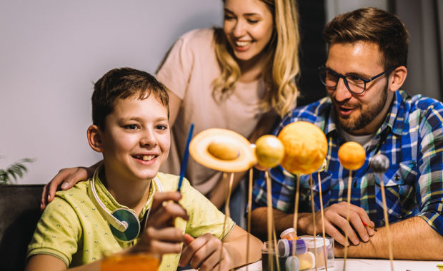 A young boy makes a solar system for a school project while his smiling parents watch.