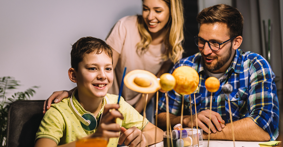 A young boy makes a solar system for a school project while his smiling parents watch.