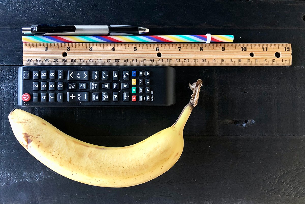 A pen, straw, TV remote control, and banana laying next to a ruler to show size differences.