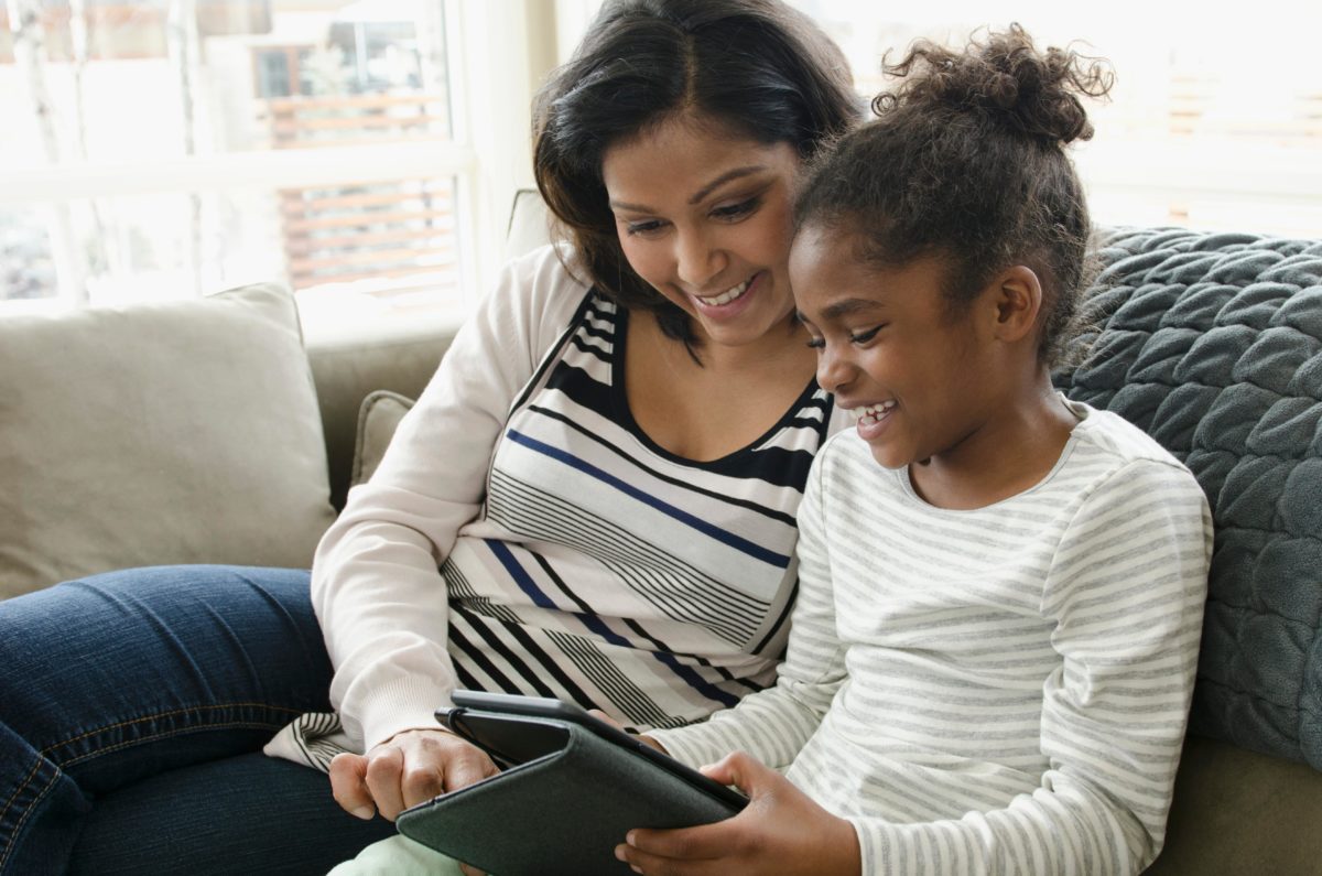 A mother and daughter use a tablet device while sitting together happily on a couch.