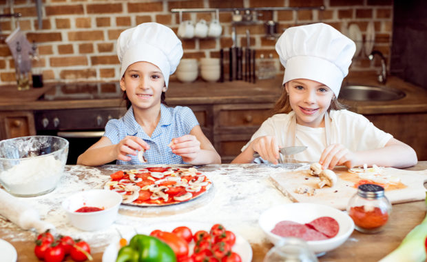 Two little girls wearing chef hats while putting toppings on a homemade pizza.