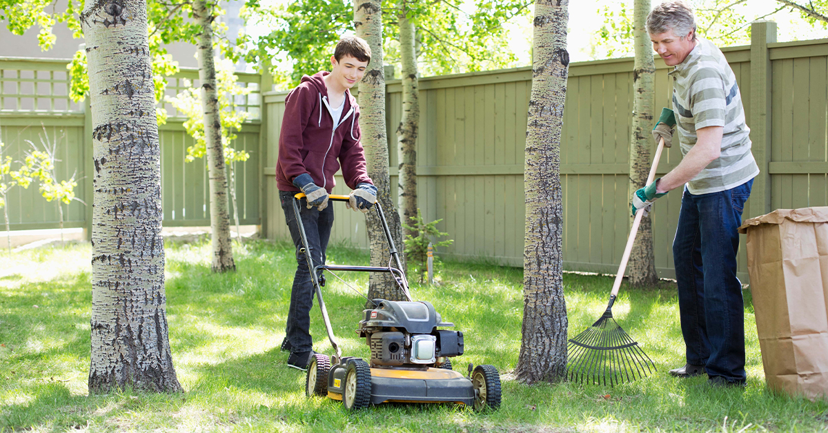 A teenage boy mows the lawn while an older man rakes.