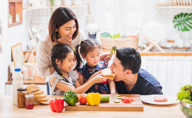 A mom, dad, and two young daughters make healthy snacks together in a kitchen.