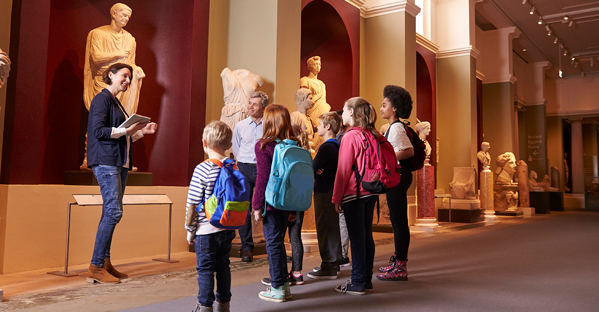 A group of students and female teacher view historic sculptures at a museum.