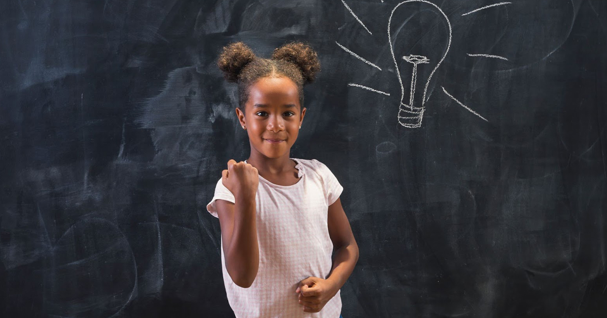 A young girl smiles confidently in front of a chalkboard.