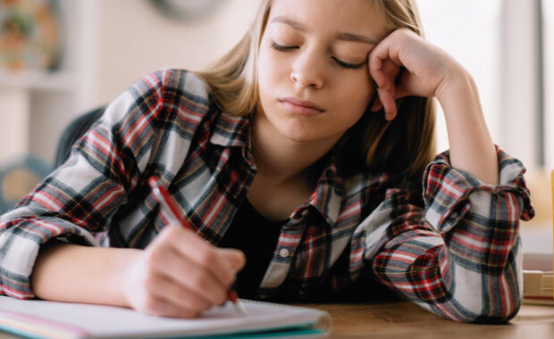 Teen girl frowns while writing in a notebook at her desk.