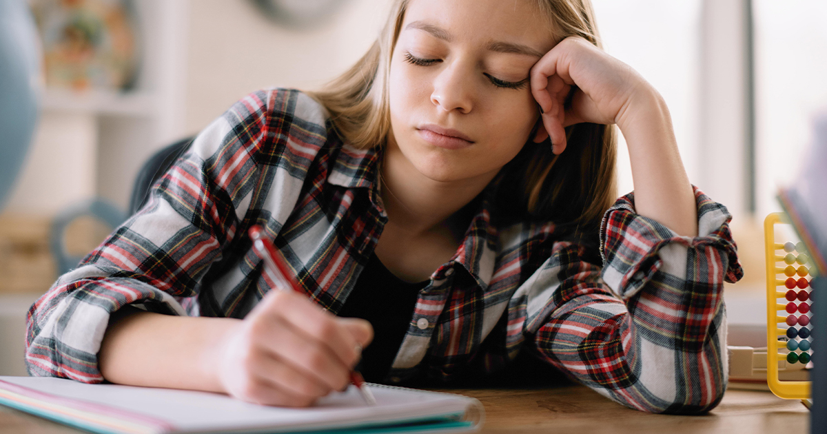 Teen girl frowns while writing in a notebook at her desk.