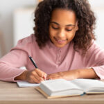 Girl writes in a notebook at a wooden desk.