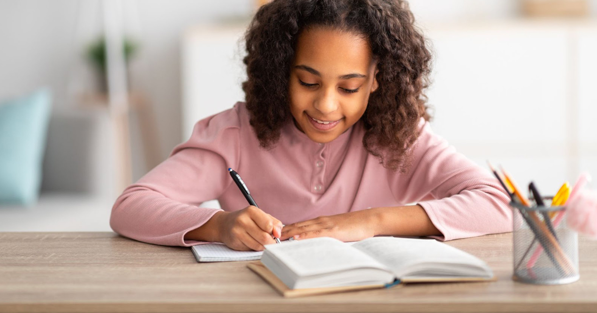 Girl writes in a notebook at a wooden desk.