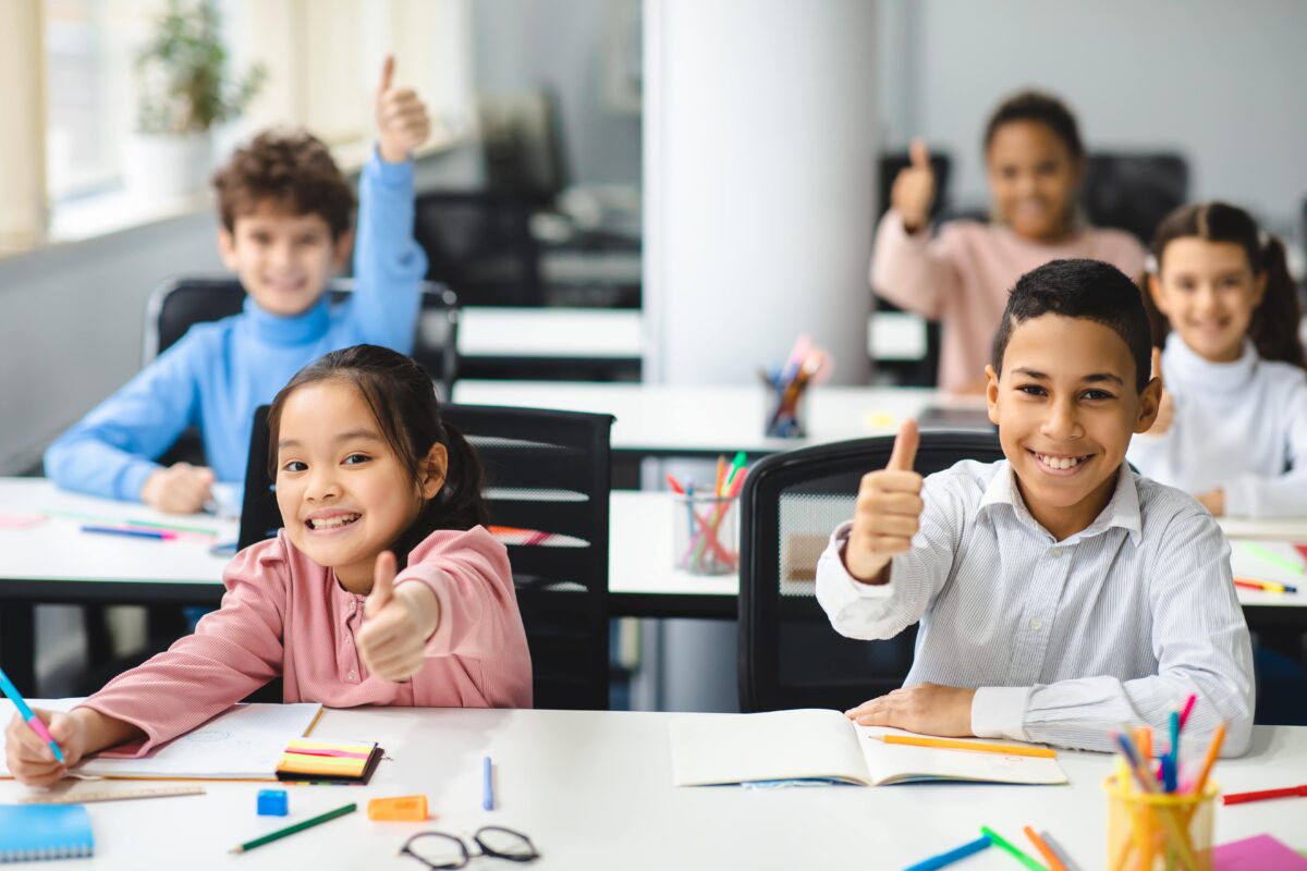 A group of students smile and give a thumbs up while working at their desks.
