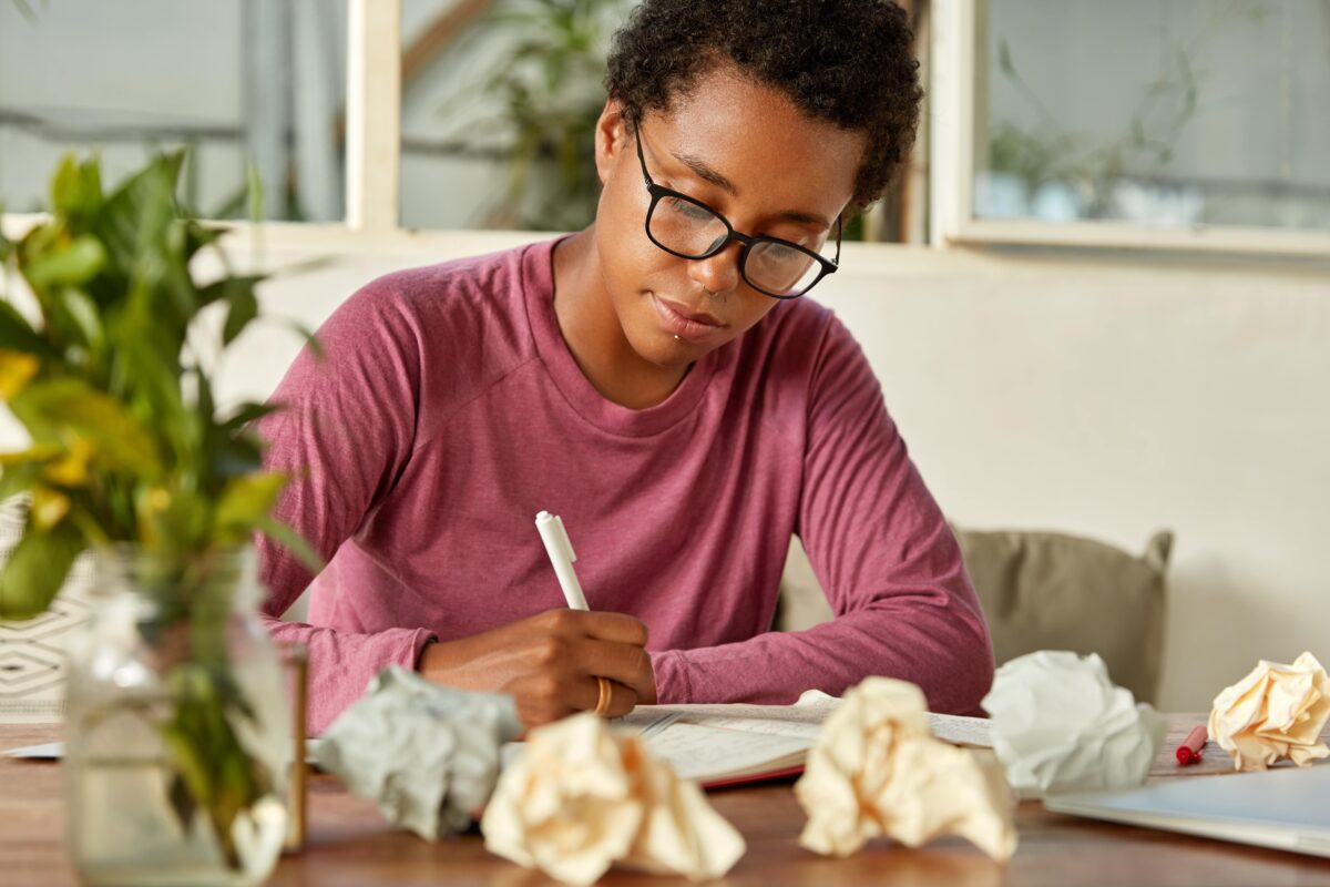 A student writes at a desk covered with crumbled up balls of paper.
