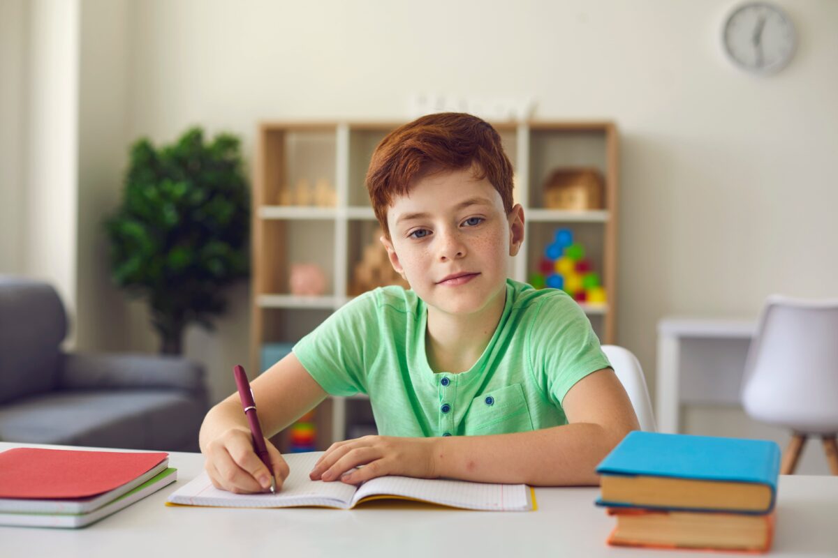 A young boy in a bright green shirt writes at his desk.