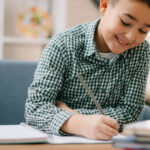 Young boy smiles while writing in a spiral notebook.