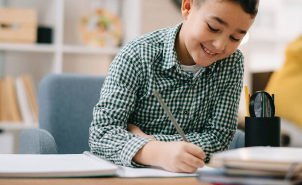 Young boy smiles while writing in a spiral notebook.
