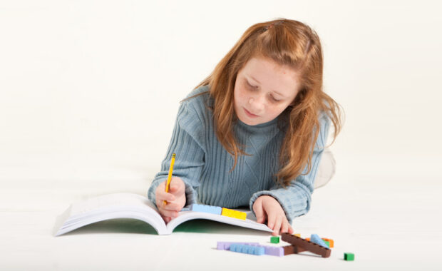 A young girl does math homework on the floor with math manipulatives beside her.