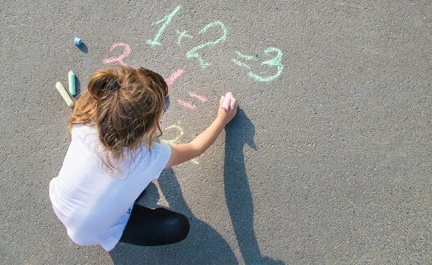 A young girl draws math facts with sidewalk chalk.