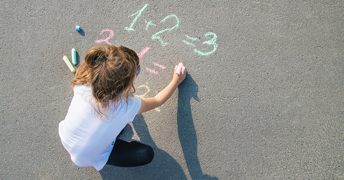 A young girl draws math facts with sidewalk chalk.